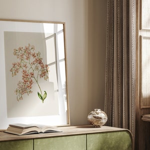 May include: A framed botanical print of a pink flower with green leaves on a light beige background. The print is hanging on a wall above a wooden table with a book and a decorative bowl.