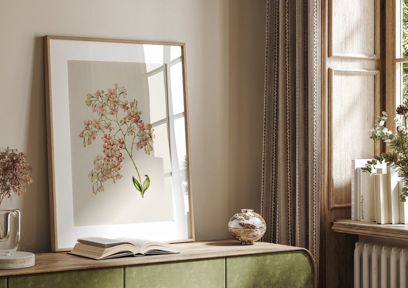 May include: A framed botanical print of a pink flower with green leaves on a light beige background. The print is hanging on a wall above a wooden table with a book and a decorative bowl.