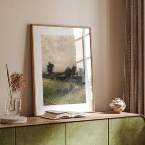 May include: A framed landscape painting of a field with trees and a cloudy sky. The painting is in a light wood frame and is hanging on a wall.