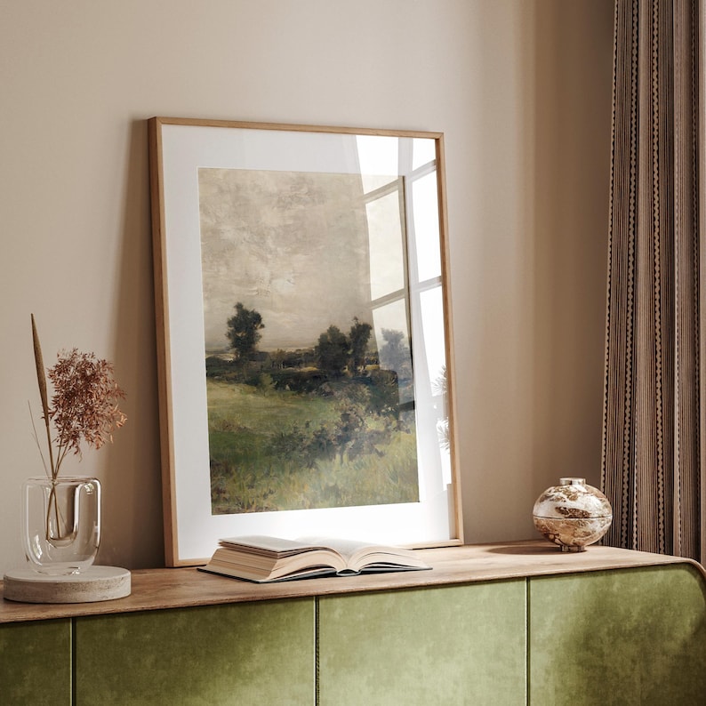 May include: A framed landscape painting of a field with trees and a cloudy sky. The painting is in a light wood frame and is hanging on a wall.