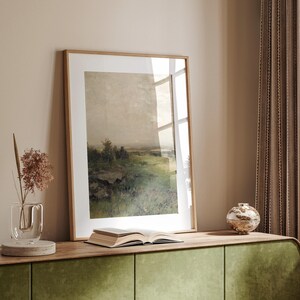 May include: A framed landscape painting of a green field with trees and a cloudy sky. The painting is hanging on a wall above a wooden shelf with a book and a decorative vase.