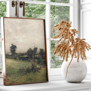 May include: A framed landscape painting of a field with trees and a cloudy sky. The painting is in a brown frame and is hanging on a white wall. A vase with dried pampas grass is in the foreground.