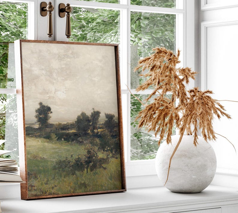 May include: A framed landscape painting of a field with trees and a cloudy sky. The painting is in a brown frame and is hanging on a white wall. A vase with dried pampas grass is in the foreground.