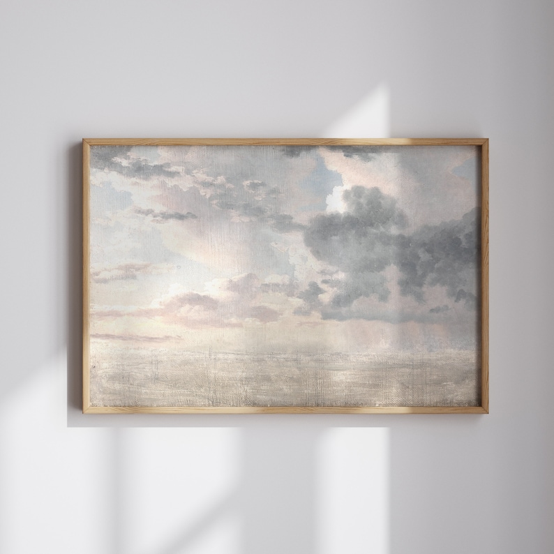 Op de afbeelding: Een ingelijste kunstdruk met een bewolkte lucht in grijstinten, blauw en roze. Het kunstwerk heeft een vintage esthetiek, gevat in een lichtbruine houten lijst. De print roept een gevoel van rust en sereniteit op.