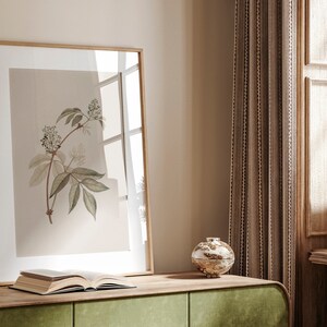 May include: A framed botanical illustration of a white flower with green leaves on a beige background. The frame is made of wood and has a light brown finish. The illustration is sitting on a wooden table with a book and a decorative object.