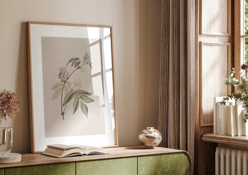May include: A framed botanical illustration of a white flower with green leaves on a beige background. The frame is made of wood and has a light brown finish. The illustration is sitting on a wooden table with a book and a decorative object.