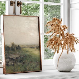 May include: A framed landscape painting of a field with trees and a cloudy sky. The painting is hanging on a white wall next to a vase with dried pampas grass.