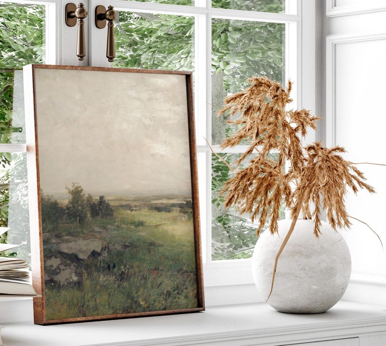 May include: A framed landscape painting of a field with trees and a cloudy sky. The painting is hanging on a white wall next to a vase with dried pampas grass.