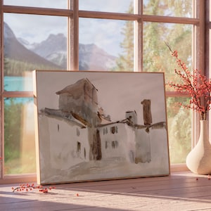 May include: A watercolor painting of a building with a brown roof and white walls, framed in a light wood frame. The painting is sitting on a wooden surface in front of a window with a view of mountains and trees.