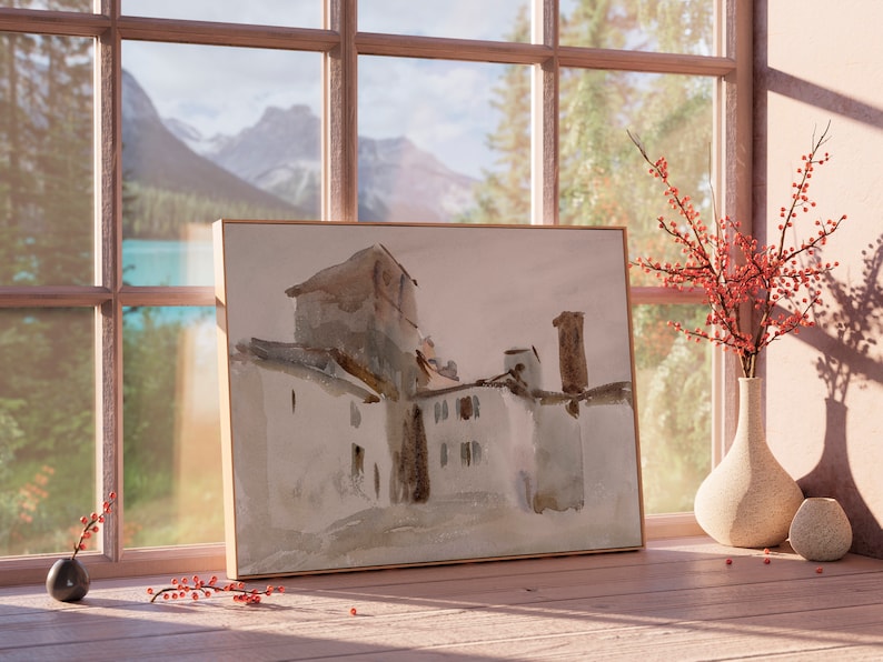 May include: A watercolor painting of a building with a brown roof and white walls, framed in a light wood frame. The painting is sitting on a wooden surface in front of a window with a view of mountains and trees.