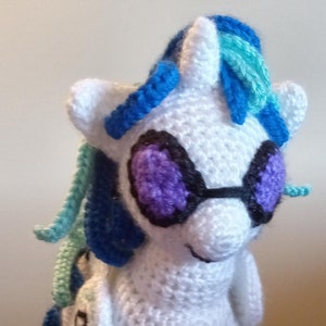 May include: A white crocheted unicorn with blue and teal yarn hair, wearing purple and black striped leg warmers and black sunglasses with purple lenses.