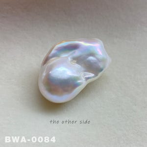 May include: A large, irregular shaped, white pearl with iridescent highlights. The pearl is smooth and has a slightly bumpy texture. The pearl is photographed on a white surface. The text "the other side" and "BWA-0084" are visible in the image.