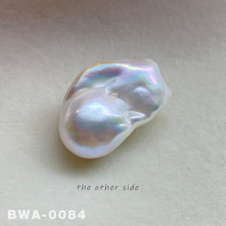 May include: A large, irregular shaped, white pearl with iridescent highlights. The pearl is smooth and has a slightly bumpy texture. The pearl is photographed on a white surface. The text "the other side" and "BWA-0084" are visible in the image.