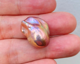 May include: A large, iridescent, baroque pearl with a pink, purple, and gold hue. The pearl is being held in a hand.