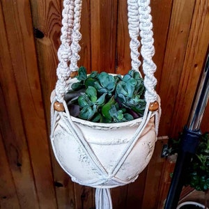 Op de afbeelding: Een witte keramische pot met een verweerde afwerking hangt aan een macramé plantenhanger met houten kralen. De pot is gevuld met een groene succulent.