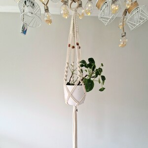 Może przedstawiać: Biały wiszący kwietnik z makramą z białą doniczką i zieloną rośliną wiszącą z białego sufitu. Kwietnik ma frędzle na dole.