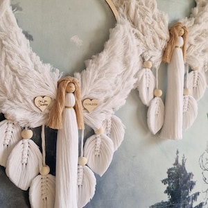 Op de afbeelding: Twee witte engelvormige macramé wandhangers met houten borden. De borden hebben de tekst "Pierwsza Komunia Swieta Nico" en "Pierwsza Rodzinny Dzien w Bigajlowie".
