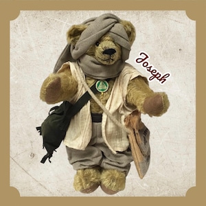 May include: A teddy bear named Joseph, wearing a turban, scarf, vest, and trousers. The bear has a green and gold pendant, a shoulder bag, and a small sack. The bear is standing and facing forward.