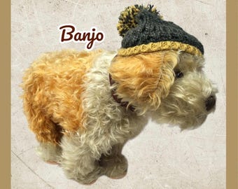 以下が含まれることがあります： クリームとタン色の毛並みを持つ犬のぬいぐるみ。濃いグレーと黄色のニット帽をかぶっています。「Banjo」の文字が茶色の筆記体で犬の上に書かれています。