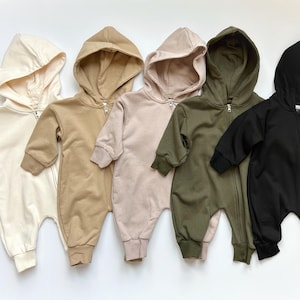 May include: Six baby rompers in a row, each with a hood and a full-length zipper. The colors include cream, tan, blush, olive green, and black. The rompers have long sleeves and are made of a soft material.