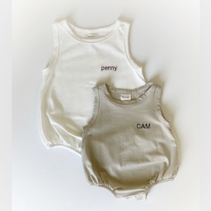 May include: Two sleeveless shirts for babies, one white and one beige. The white shirt has the name "penny" embroidered on it. The beige shirt has the name "CAM" embroidered on it.