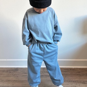 May include: A child wearing a blue sweatshirt and matching sweatpants with a black beanie. The child is wearing white sneakers with black stripes.