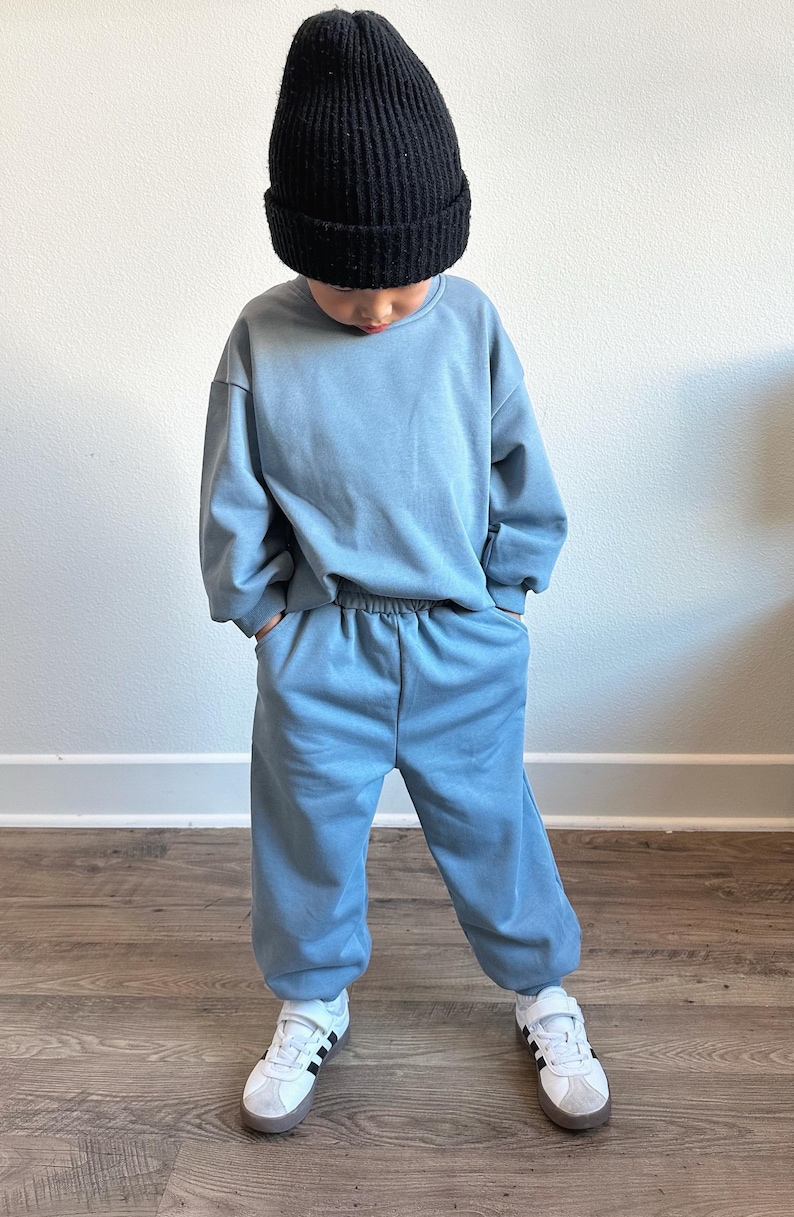 May include: A child wearing a blue sweatshirt and matching sweatpants with a black beanie. The child is wearing white sneakers with black stripes.