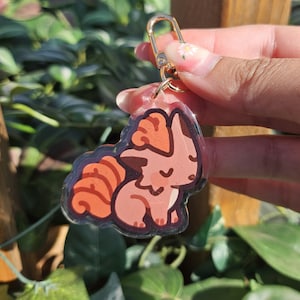 May include: A clear acrylic keychain featuring a cartoon fox-like creature in shades of peach and orange. The keychain has a gold-colored clasp and a small ring for attaching to keys or bags. The design is outlined in a dark brown.