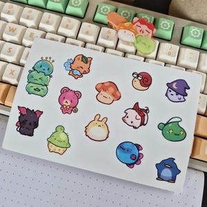 Könnte beinhalten: Ein weißer Aufkleberbogen mit verschiedenen bunten Cartoon-Figuren, darunter ein Stapel grüner Wesen, ein rosa Bär, ein Pilz, eine Schnecke, ein violetter Zauberer, ein schwarzes fledermausartiges Wesen, ein Kaktus, ein Hase, ein Schwein und ein blauer Pinguin.
