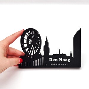 May include: Black silhouette of the Den Haag skyline, featuring a Ferris wheel, buildings, and the city name in white text.