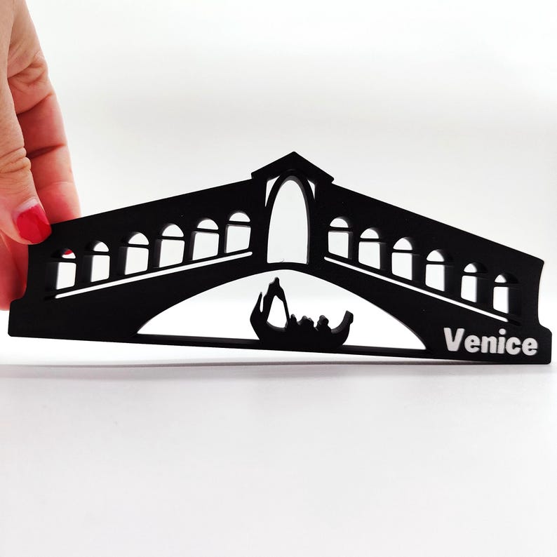 May include: Black silhouette of the Rialto Bridge in Venice, Italy, with a gondola underneath. The bridge has arches and the word "Venice" is printed on the bottom right.