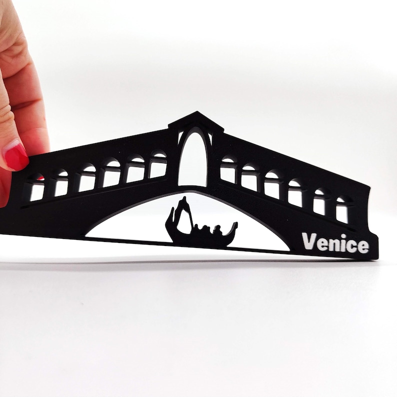 May include: Black silhouette of the Rialto Bridge in Venice, Italy, with a gondola underneath. The bridge has a large arch in the centre and smaller arches on either side. The word "Venice" is printed in white on the bottom right corner.