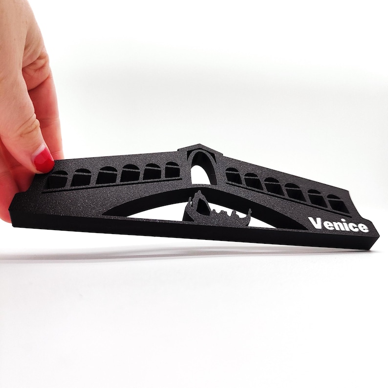 May include: Black foam cutout of the Rialto Bridge in Venice, Italy. The bridge is arched and has a gondola underneath. The word "Venice" is printed in white on the right side of the cutout.