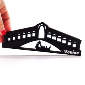 May include: Black silhouette of the Rialto Bridge in Venice, Italy. The bridge is made of wood and has a gondola underneath it. The word "Venice" is written in white letters on the bottom right of the bridge.