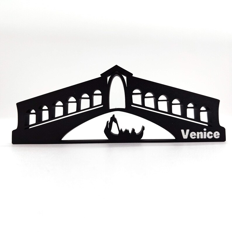 May include: Black silhouette of the Rialto Bridge in Venice, Italy. The bridge has a large archway and many smaller arches. A gondola is visible under the bridge. The word "Venice" is written in white below the bridge.