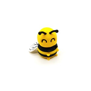 May include: A yellow and black bee figurine with a smiling face and small wings. The bee has black stripes around its body and small yellow legs. The wings are white with black detailing. The figurine is set against a white background.