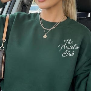 Może przedstawiać: Ciemnozielona bluza z okrągłym dekoltem i białym napisem The Matcha Club wyhaftowanym kursywą na lewej piersi. Odzież posiada długie rękawy z prążkowanymi mankietami oraz pasujący prążkowany dekolt, co nadaje jej prosty i klasyczny wygląd.