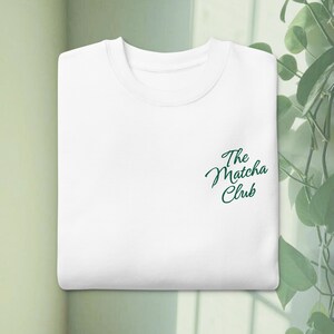 Może przedstawiać: Złożona biała bluza z okrągłym dekoltem i napisem The Matcha Club wyhaftowanym ciemnozieloną kursywą po prawej stronie klatki piersiowej. Odzież posiada prążkowany kołnierzyk oraz prosty kr&oacute;j z długimi rękawami złożonymi do wewnątrz.