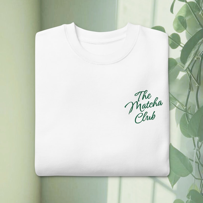 Może przedstawiać: Złożona biała bluza z okrągłym dekoltem i napisem The Matcha Club wyhaftowanym ciemnozieloną kursywą po prawej stronie klatki piersiowej. Odzież posiada prążkowany kołnierzyk oraz prosty kr&oacute;j z długimi rękawami złożonymi do wewnątrz.