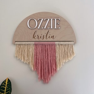 May include: A wooden half-circle sign with the names "Ozzie" and "kristin" written in white letters. The sign is hanging from a macrame wall hanging with cream and pink yarn.