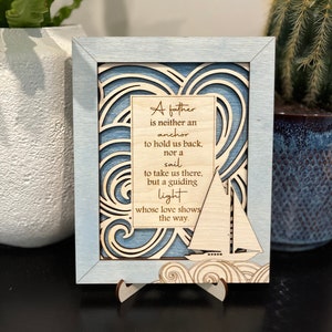 Op de afbeelding: Een houten bord met een blauw en wit golvend ontwerp en een citaat over een vader die een leidende licht is. Het citaat luidt: "Een vader is noch een anker om ons terug te houden, noch een zeil om ons daarheen te brengen, maar een leidende licht wiens liefde de weg wijst."