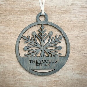 May include: A grey wooden Christmas ornament featuring a snowflake design within a circular frame. It has a white ribbon for hanging and the text "THE SCOTTS EST. 2016" is engraved at the bottom. The ornament is displayed on a light wood surface.