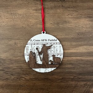 May include: A round wooden ornament with a white background and a brown silhouette of the nativity scene. The ornament features the words "O, Come All Ye Faithful" and music notes.