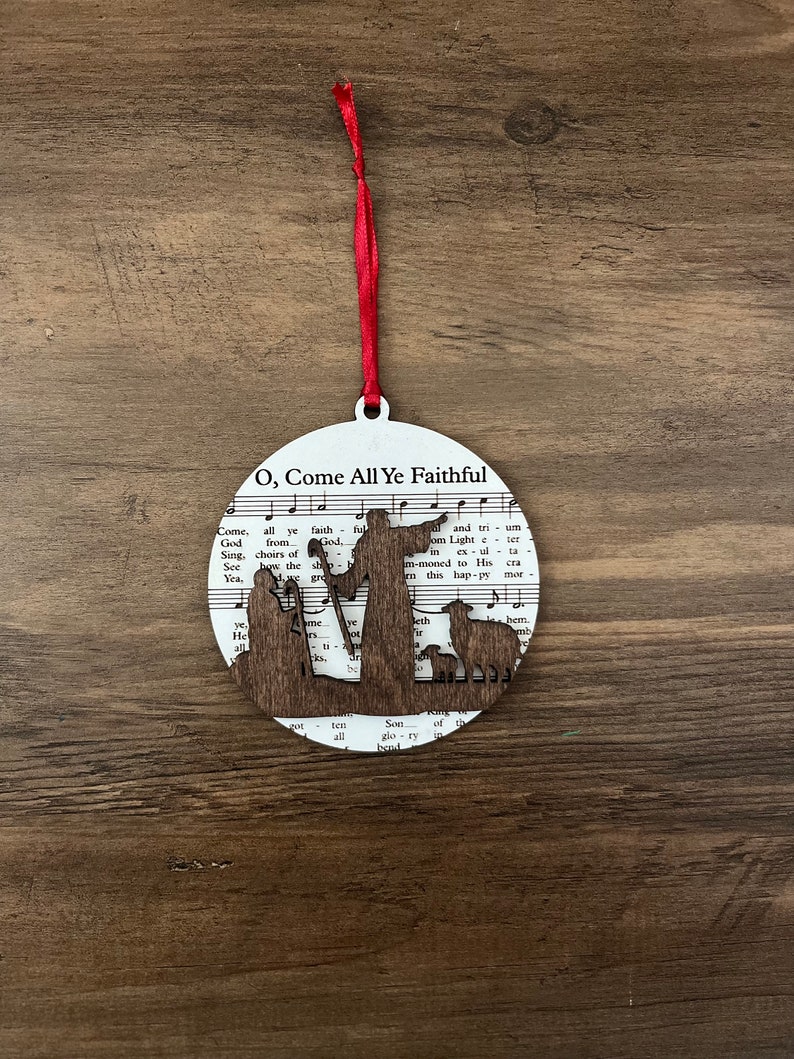 May include: A round wooden ornament with a white background and a brown silhouette of the nativity scene. The ornament features the words "O, Come All Ye Faithful" and music notes.