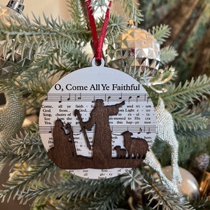 May include: A wooden Christmas ornament featuring a silhouette of the nativity scene with the words "O, Come All Ye Faithful" and music notes printed on the background.