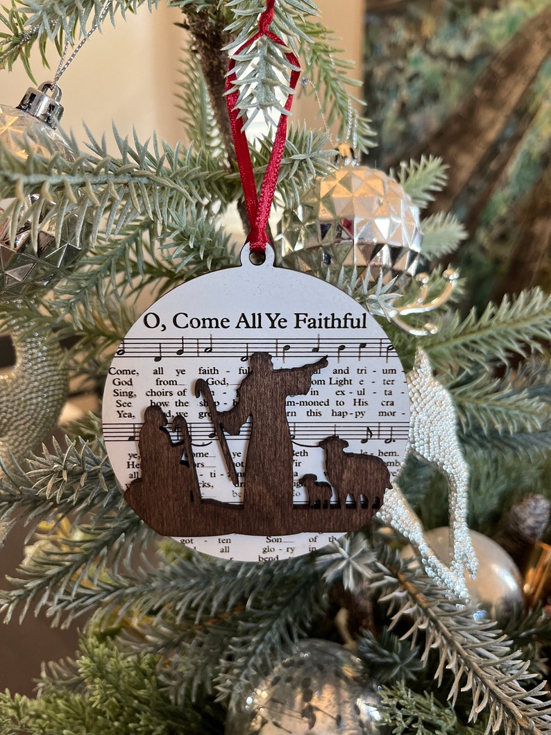 May include: A wooden Christmas ornament featuring a silhouette of the nativity scene with the words "O, Come All Ye Faithful" and music notes printed on the background.