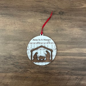 May include: White Christmas ornament with a red ribbon. The ornament features a silhouette of the nativity scene with the text "Away In A Manger" printed on the front.