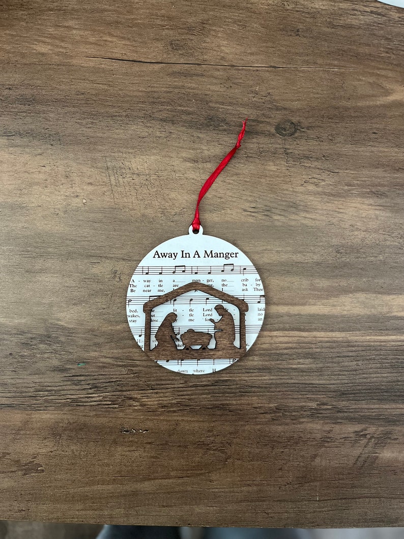 May include: White Christmas ornament with a red ribbon. The ornament features a silhouette of the nativity scene with the text "Away In A Manger" printed on the front.