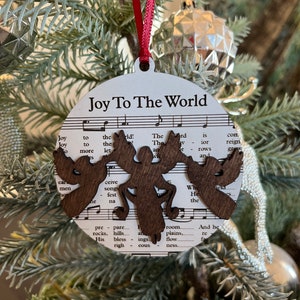May include: A white round Christmas ornament with the words "Joy To The World" printed on it. The ornament features a brown wooden cutout of a stylized angel with wings and a halo. The ornament is decorated with sheet music in the background.