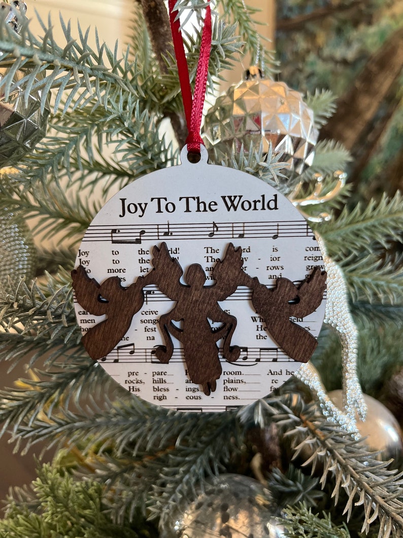 May include: A white round Christmas ornament with the words "Joy To The World" printed on it. The ornament features a brown wooden cutout of a stylized angel with wings and a halo. The ornament is decorated with sheet music in the background.